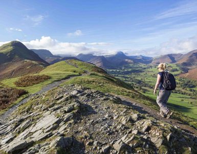 Daffodil | Woman hiking on a rocky hilltop path overlooking rolling green valleys and distant peaks in the Lake District on a clear, sunny day. ?? Daffodil